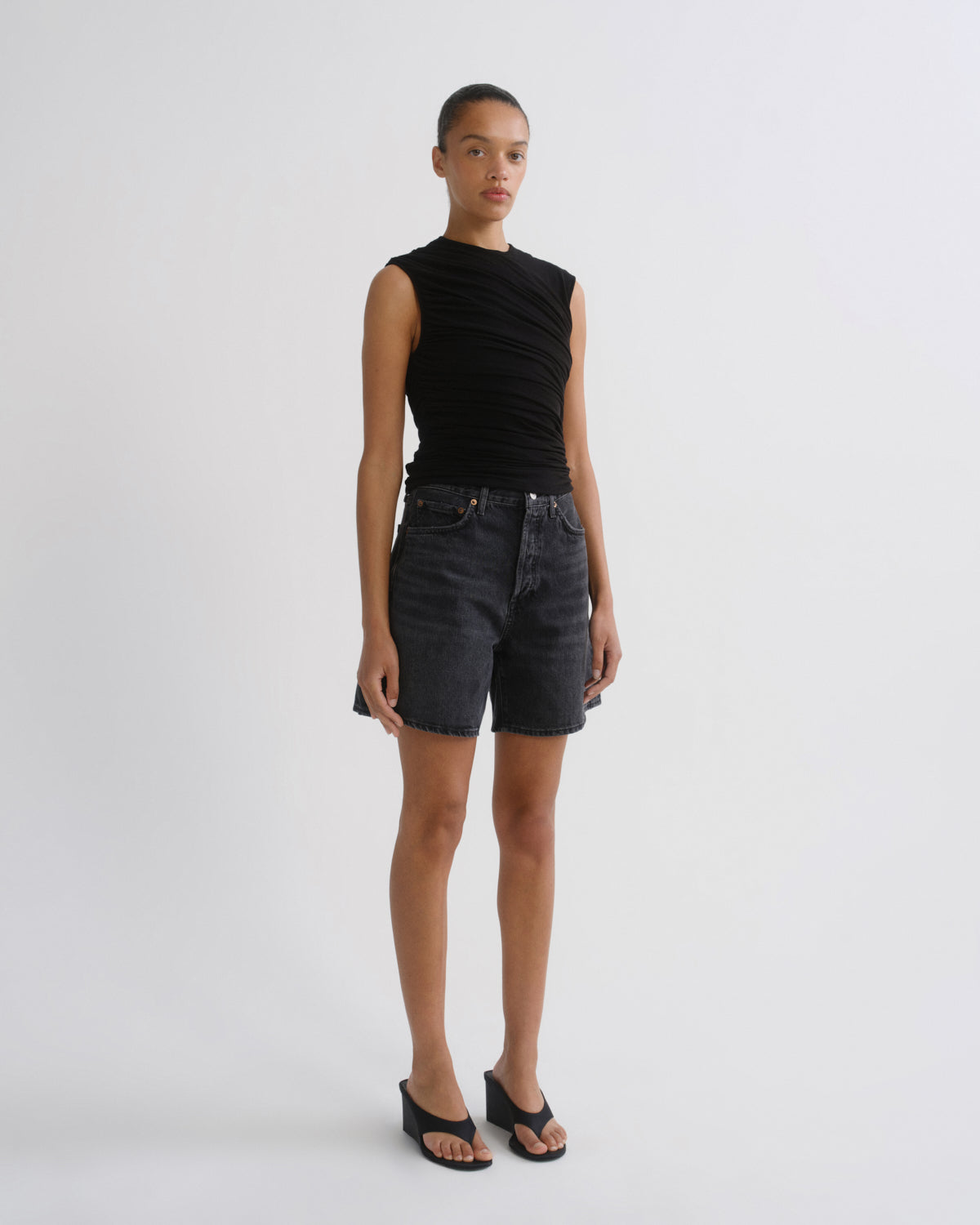 A model stands against a light gray studio background wearing a sleeveless black ruched top, dark gray denim shorts, and black wedge sandals.