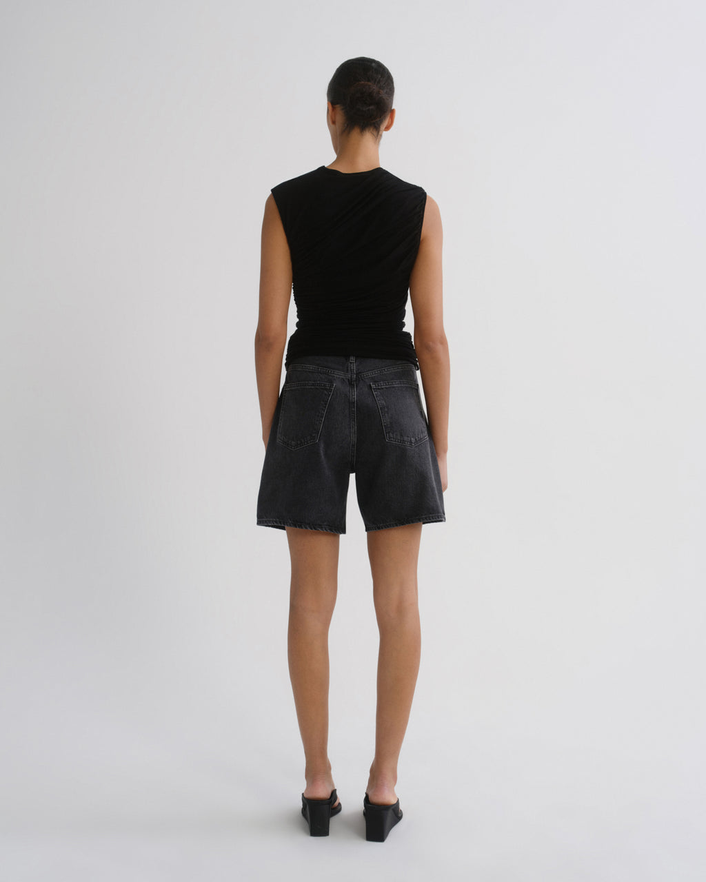 A model viewed from the back wearing a black sleeveless top with a draped neckline and dark washed denim shorts, standing against a light gray studio background.