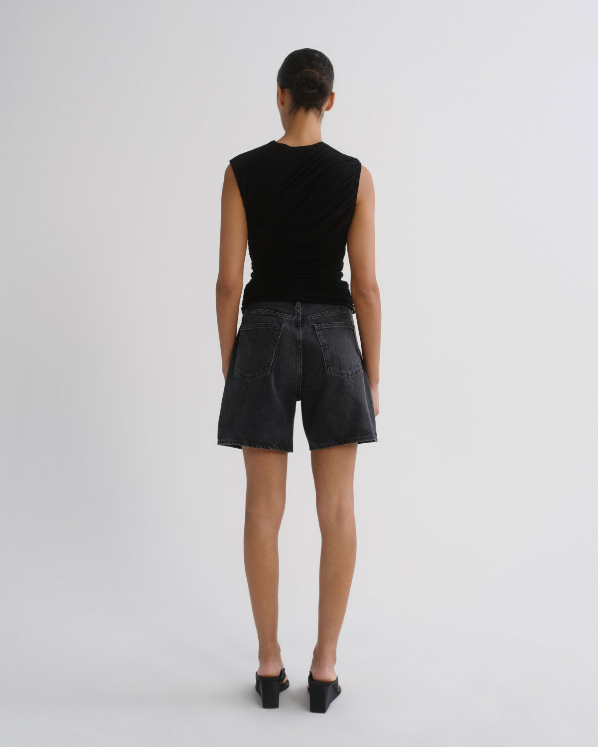 A model viewed from the back wearing a black sleeveless top with a draped neckline and dark washed denim shorts, standing against a light gray studio background.