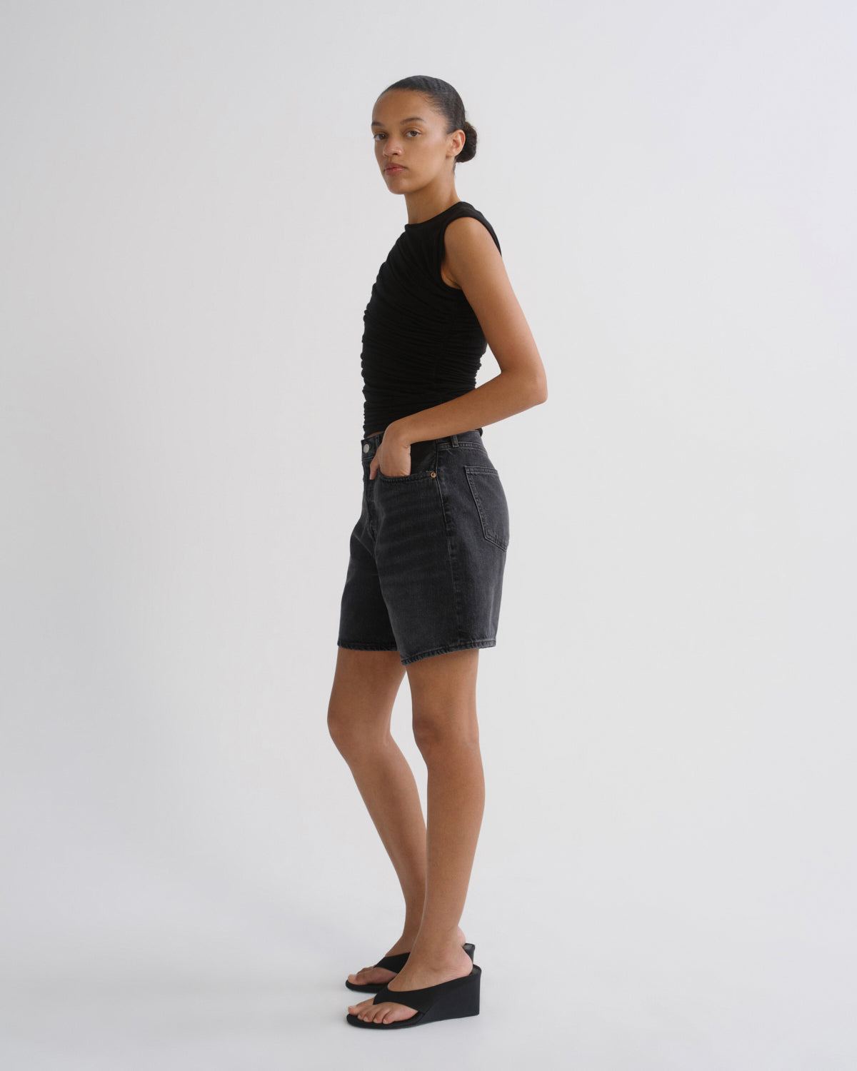 A model stands in profile wearing a black sleeveless top, dark gray denim shorts, and black wedge sandals against a plain light gray studio background.