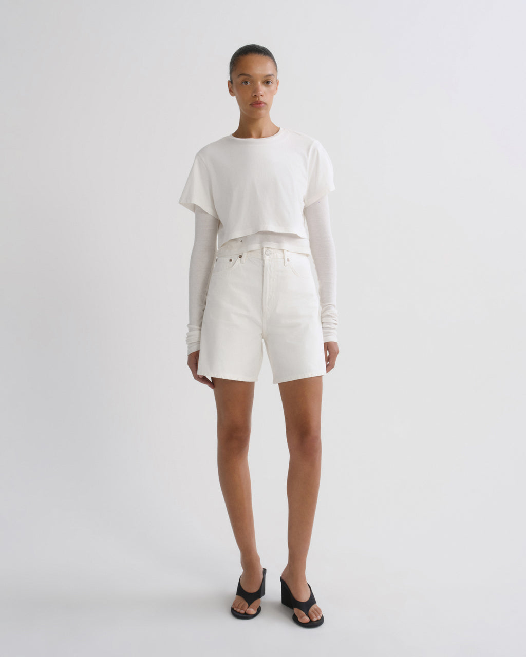 A model stands against a light gray studio background wearing a white cropped short-sleeve top over a long-sleeve white shirt, white shorts, and black slide sandals.