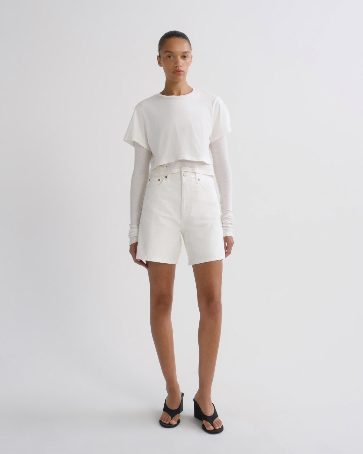 A model stands against a light gray studio background wearing a white cropped short-sleeve top over a long-sleeve white shirt, white shorts, and black slide sandals.