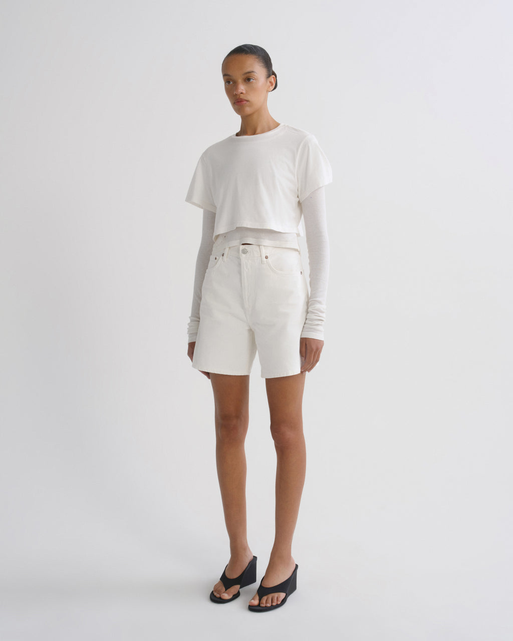 A model stands against a light gray studio background wearing a white cropped long-sleeve t-shirt, white shorts, and black slide sandals.