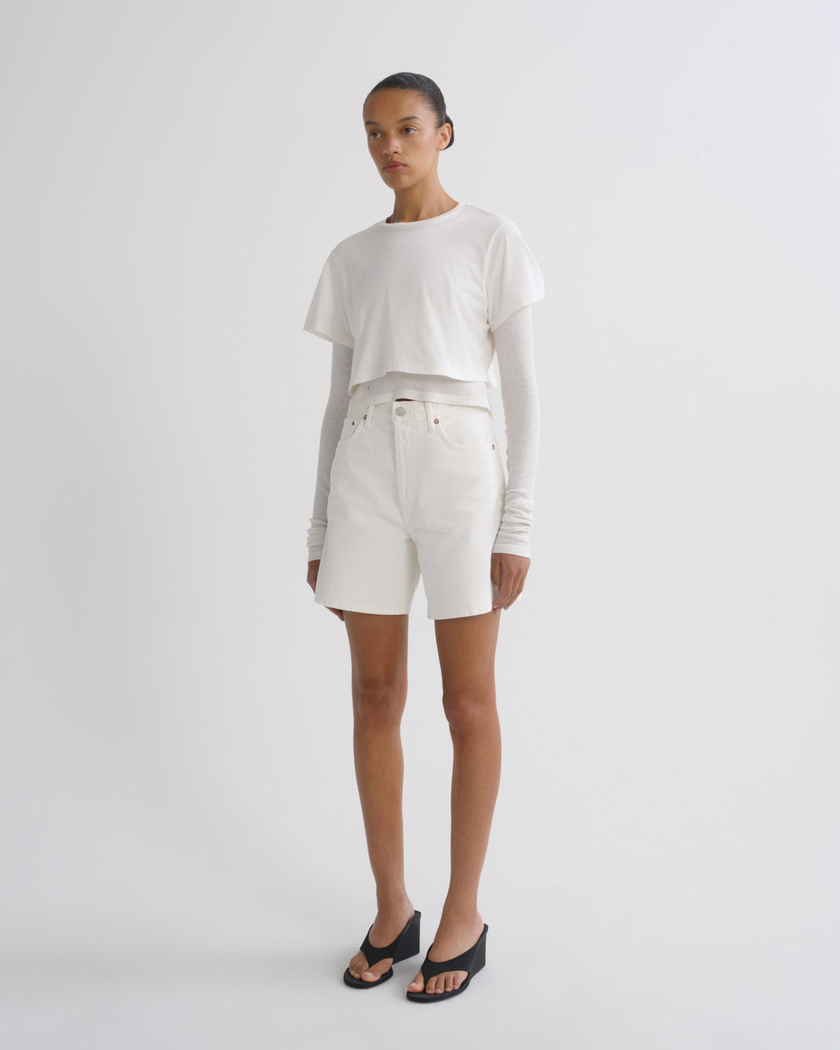 A model stands against a light gray studio background wearing a white cropped long-sleeve t-shirt, white shorts, and black slide sandals.