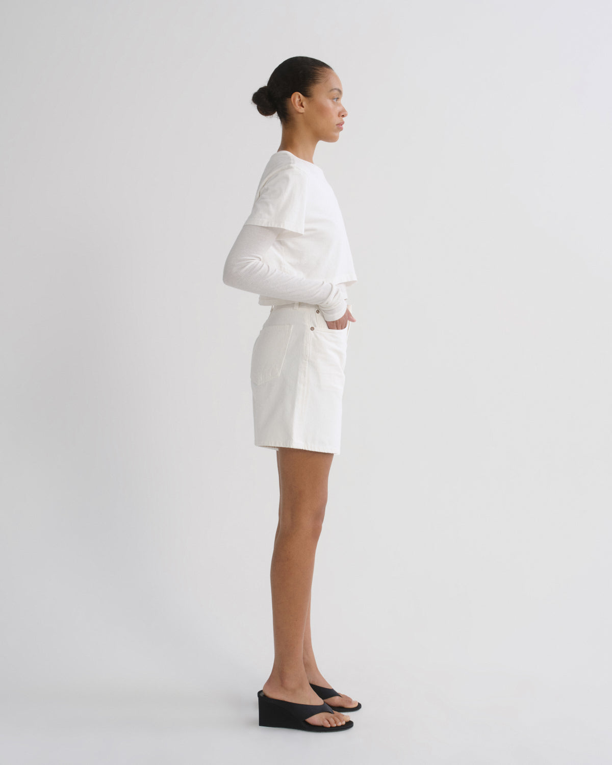 A model is shown in profile wearing a white long-sleeve top and matching white shorts, paired with black wedge sandals, against a plain light gray studio background.