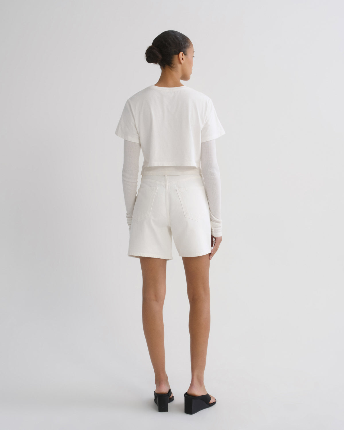 A model viewed from the back wearing a white cropped short-sleeve t-shirt over a long-sleeve white top, paired with white shorts and black wedge sandals, against a light gray studio background.