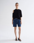 A model stands against a light gray studio background wearing a black cold-shoulder t-shirt, dark blue denim shorts, and black wedge sandals.
