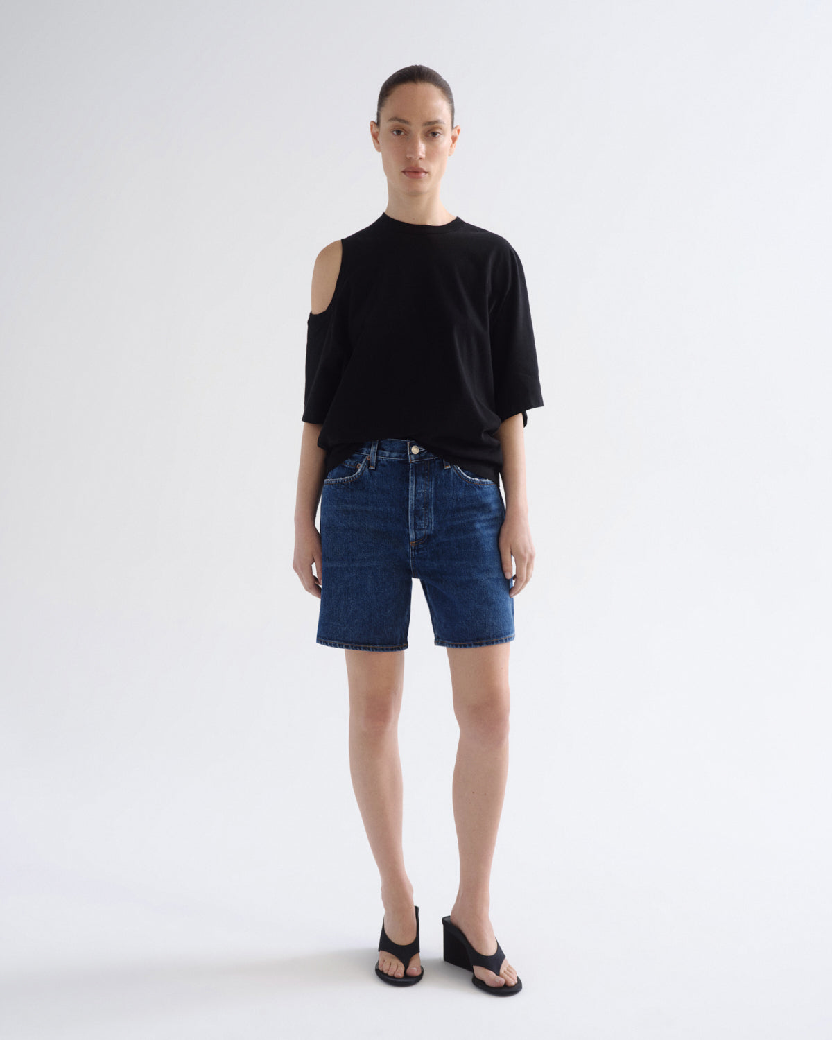 A model stands against a light gray studio background wearing a black cold-shoulder t-shirt, dark blue denim shorts, and black wedge sandals.
