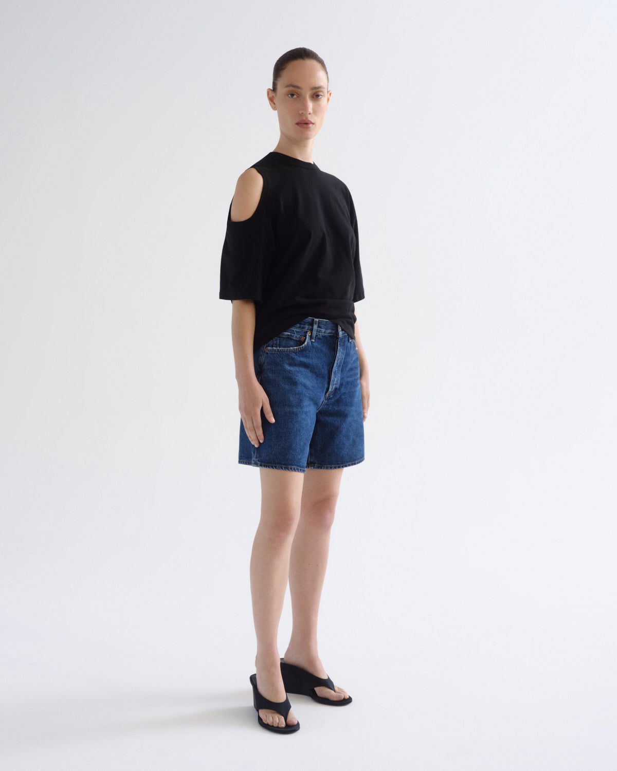 A model stands in profile wearing a black cold-shoulder short-sleeve top, blue denim shorts, and black sandals against a plain light gray studio background.