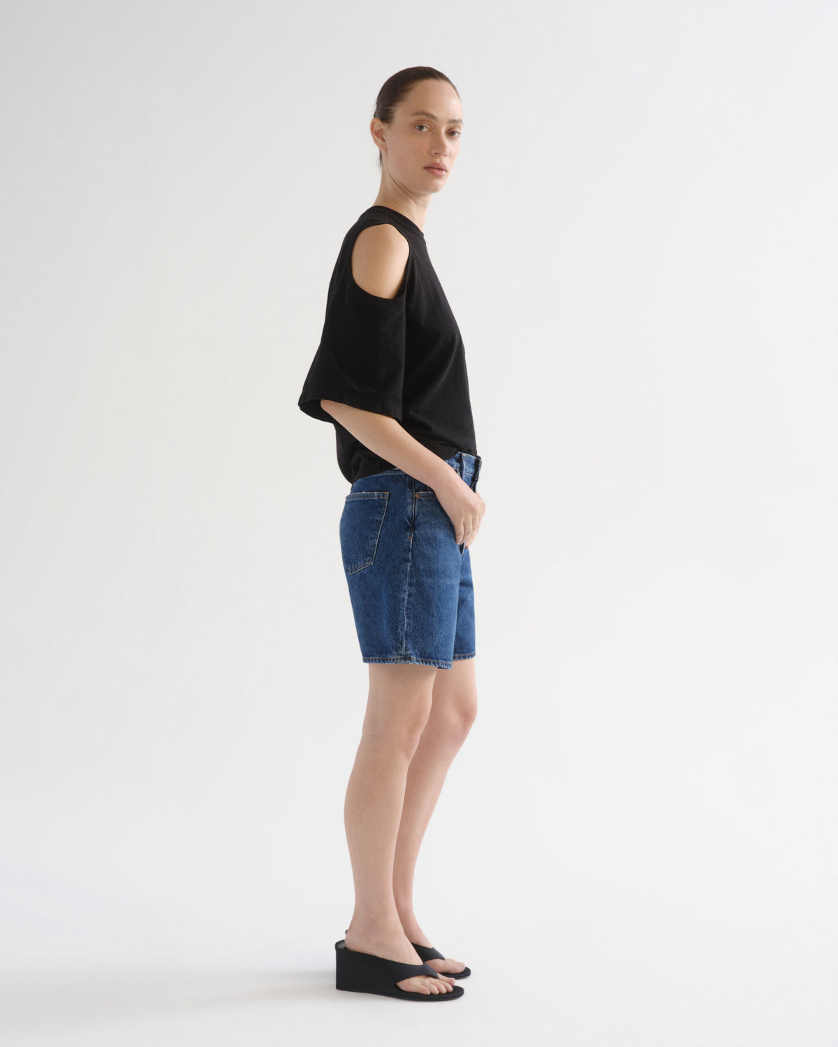 A model stands in profile wearing a black cold-shoulder knit top, blue denim shorts, and black wedge sandals against a plain white studio background.