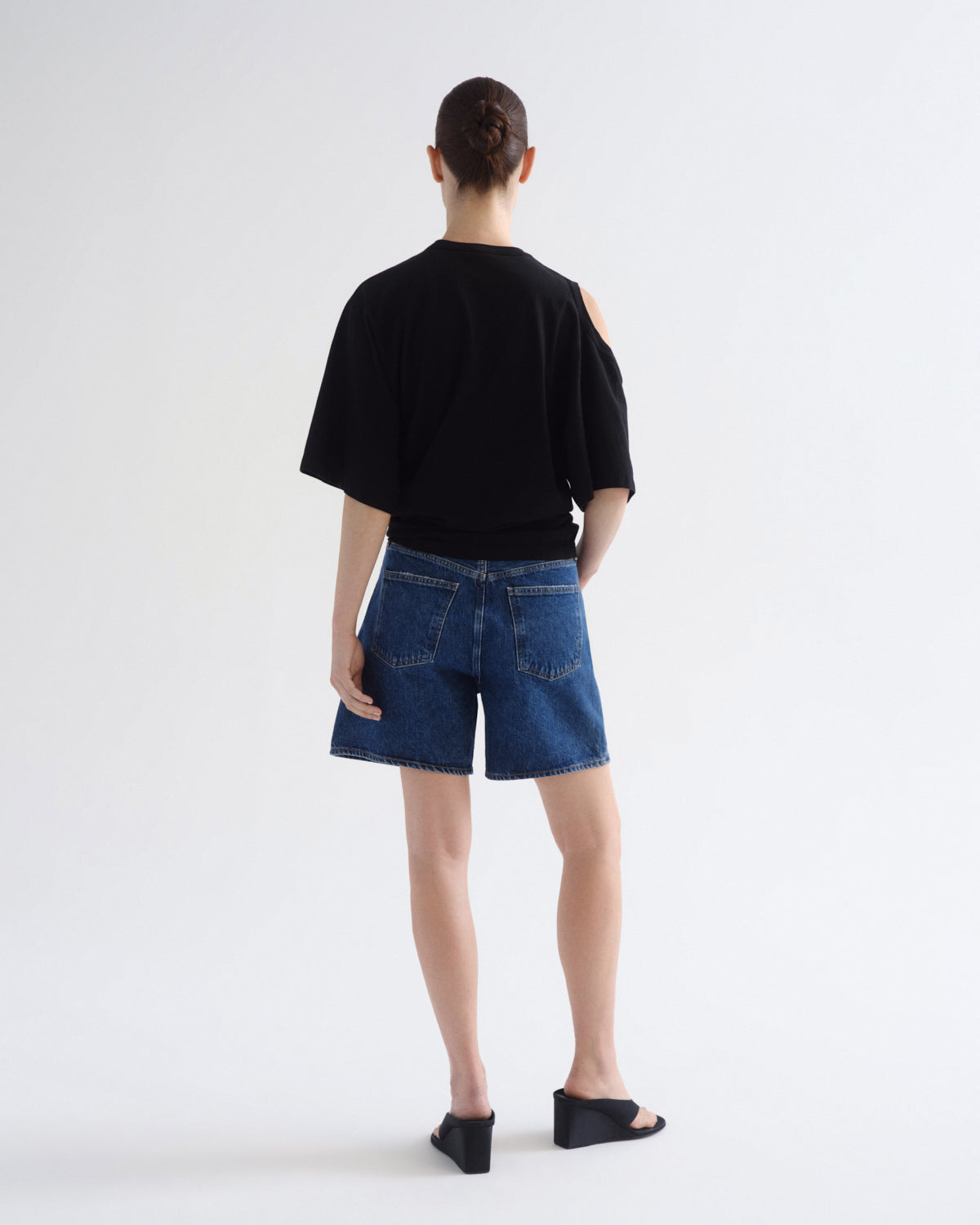 A model shown from the back wearing a black short-sleeve t-shirt with a shoulder cutout, blue denim shorts, and black wedge sandals, set against a plain white studio background.