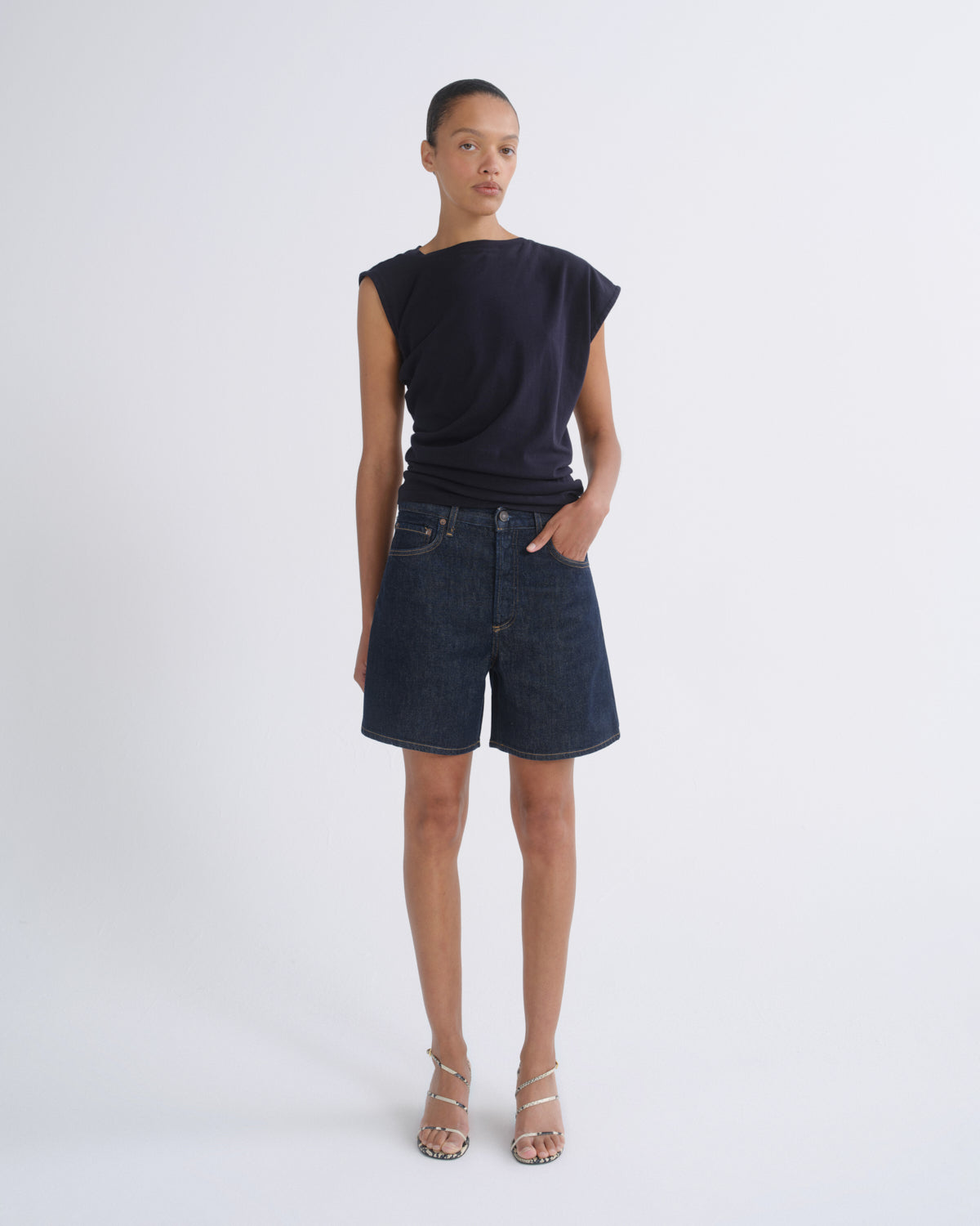A model stands against a light gray studio background wearing a dark navy sleeveless top, dark blue denim shorts, and beige strappy high-heeled sandals.