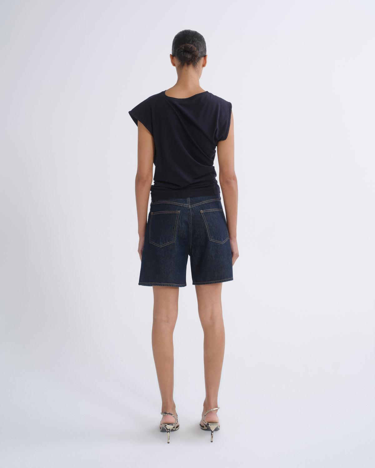 A model viewed from behind wearing a dark navy draped short-sleeve top, dark blue denim shorts, and metallic high-heeled sandals against a light gray studio background.