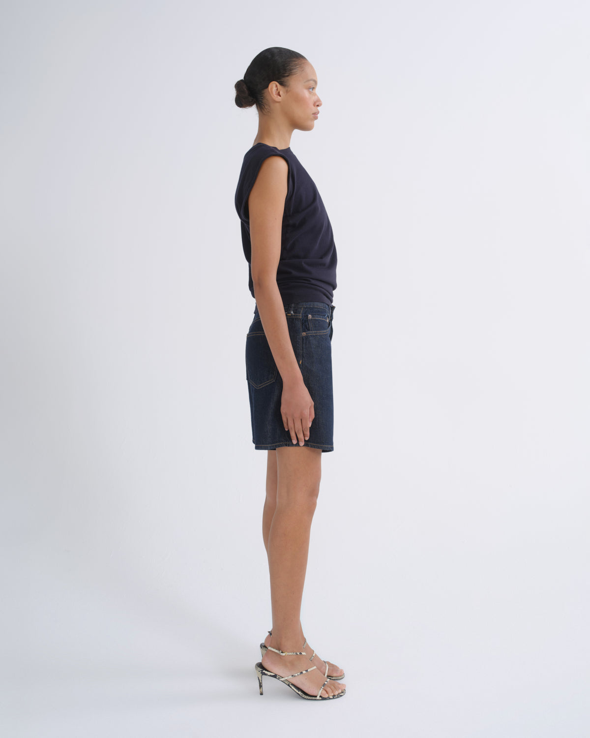 A model stands in profile wearing a dark navy sleeveless top, a dark blue denim mini skirt, and metallic silver strappy high-heeled sandals against a plain light gray studio background.