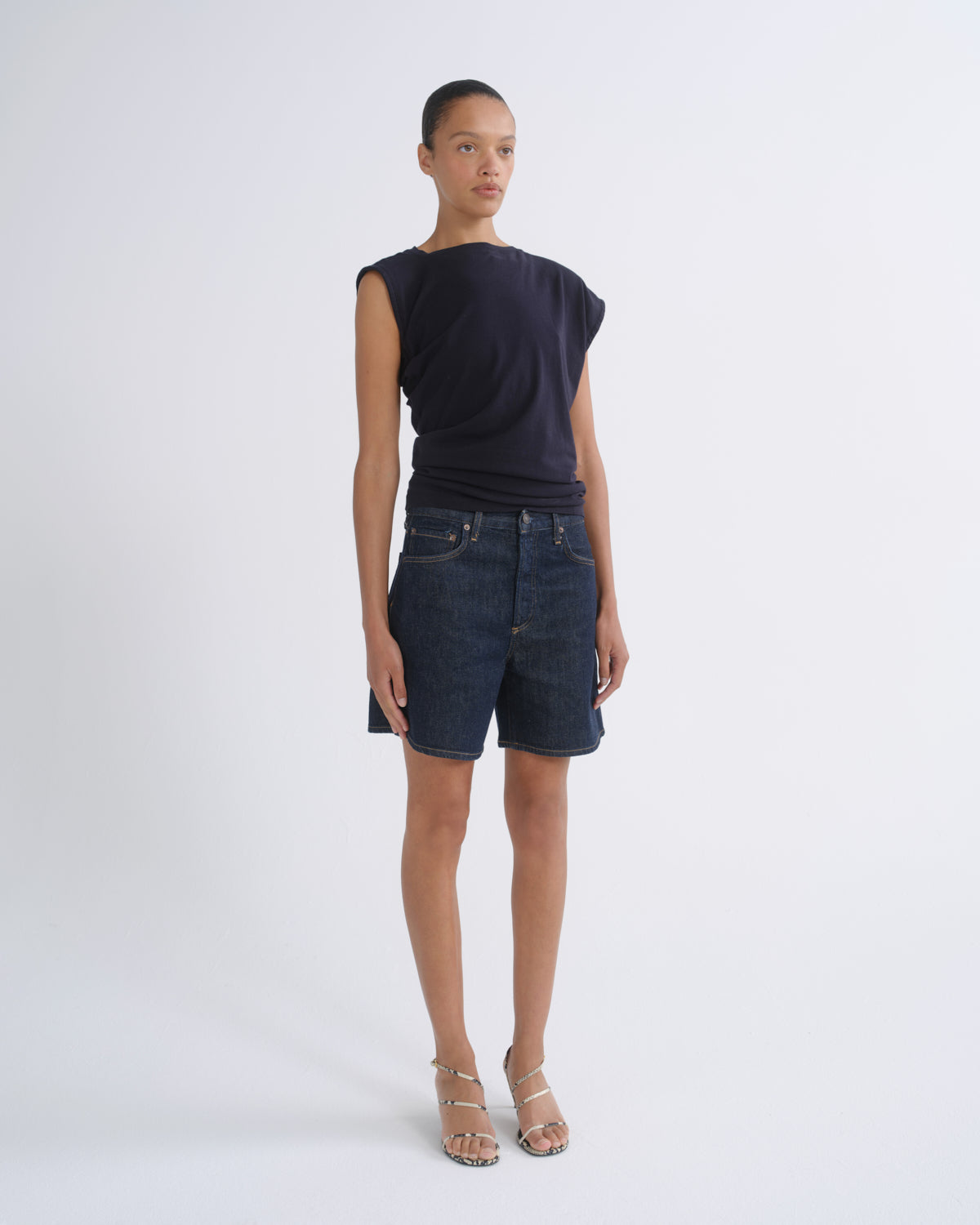 A model stands against a light gray studio background wearing a dark navy sleeveless top, dark blue denim shorts, and beige strappy sandals.