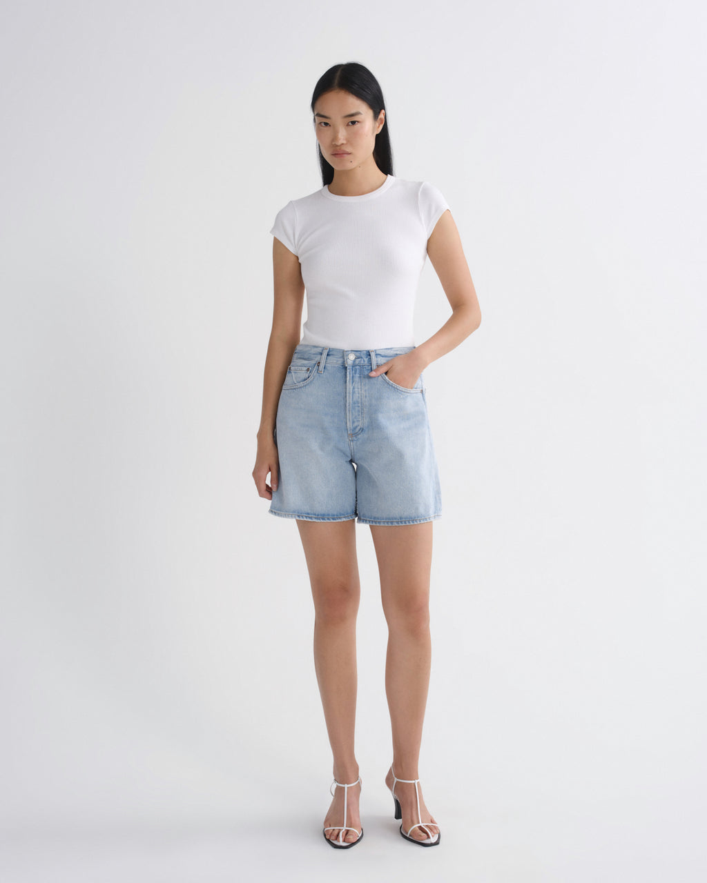 A woman modeling a white short-sleeve crewneck t-shirt and light-wash denim shorts against a plain light gray studio background.