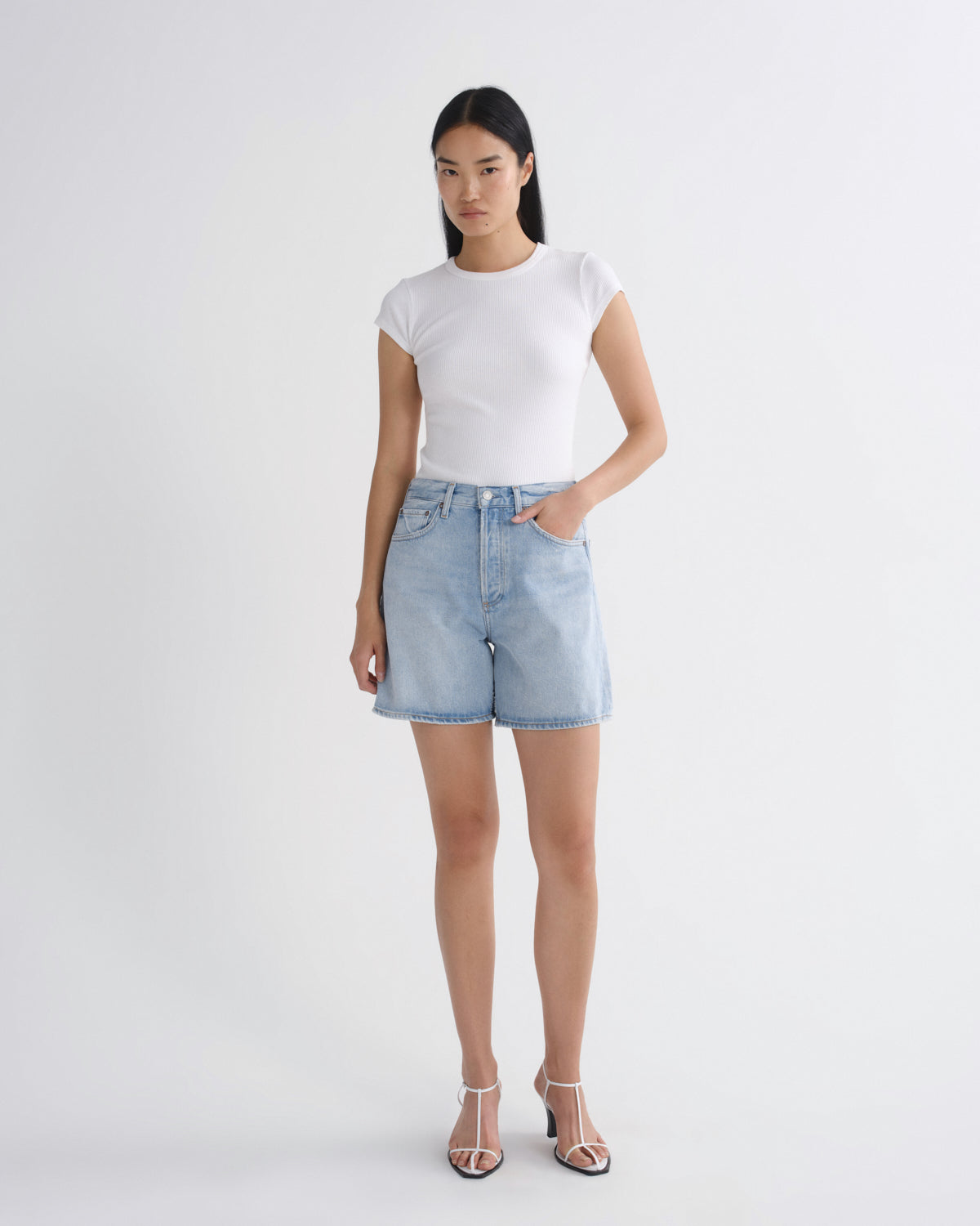 A woman modeling a white short-sleeve crewneck t-shirt and light-wash denim shorts against a plain light gray studio background.