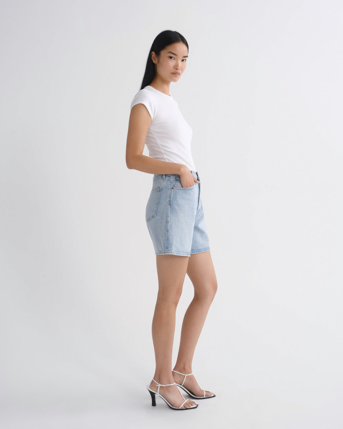 A woman modeling a white short-sleeve crewneck t-shirt and light wash denim shorts, paired with white strappy high-heeled sandals, against a plain light gray studio background.