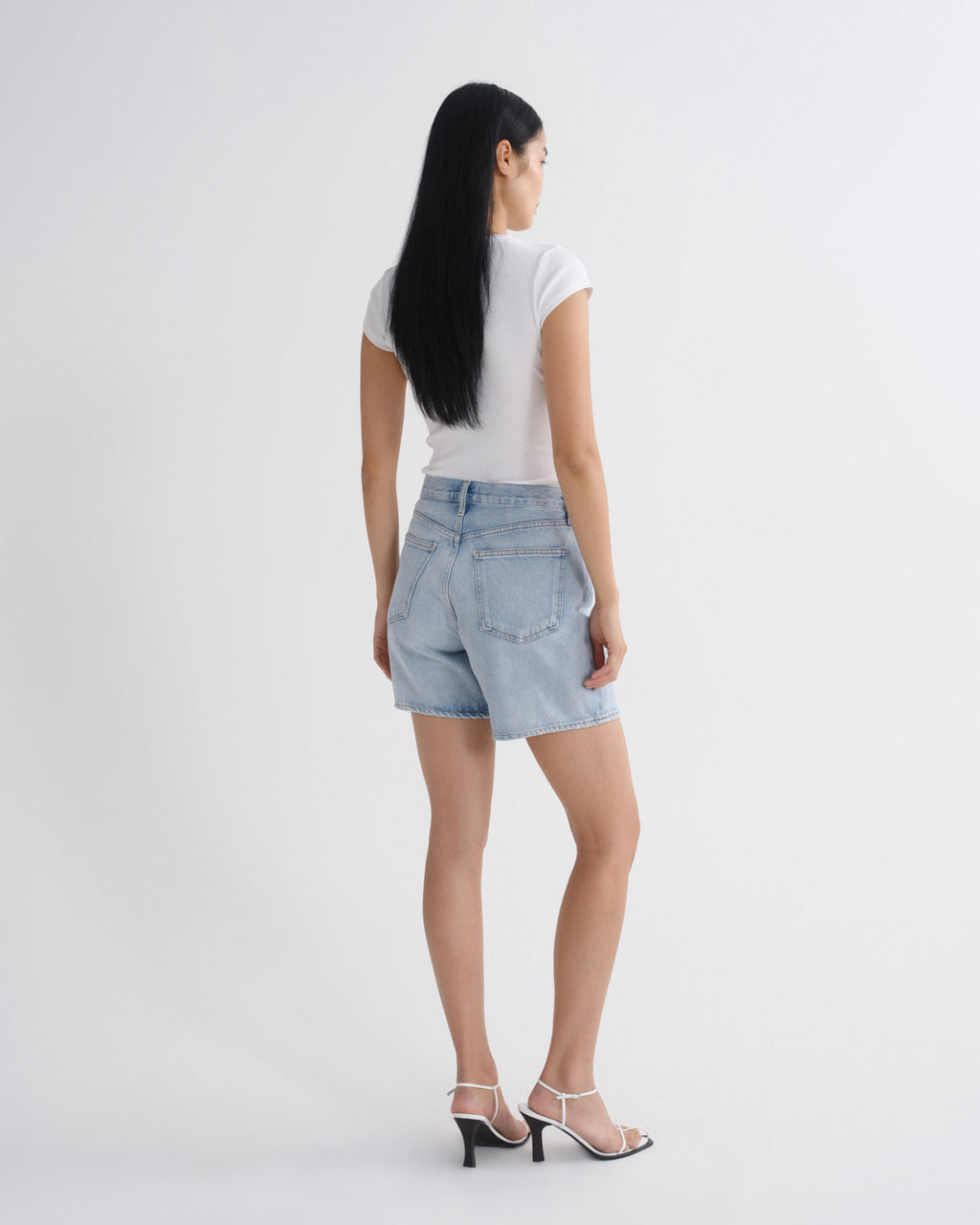 A woman shown from the back wearing light-wash denim shorts, a white short-sleeve top, and white heeled sandals against a plain light gray studio background.