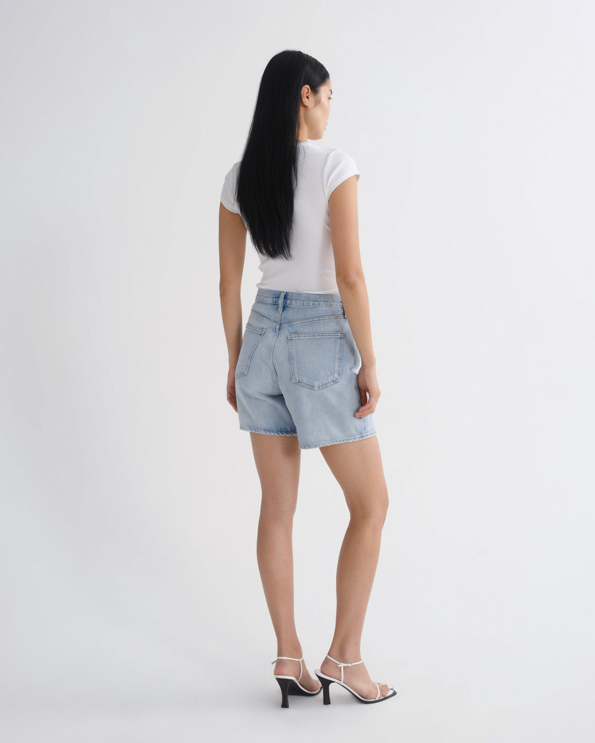 A woman shown from the back wearing light-wash denim shorts, a white short-sleeve top, and white heeled sandals against a plain light gray studio background.