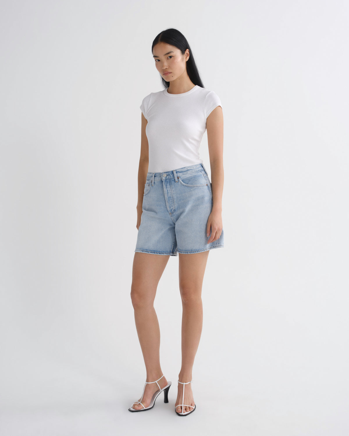 A woman modeling a white short-sleeve crewneck t-shirt and light-wash blue denim shorts, paired with clear-strap high-heeled sandals, against a plain white studio background.