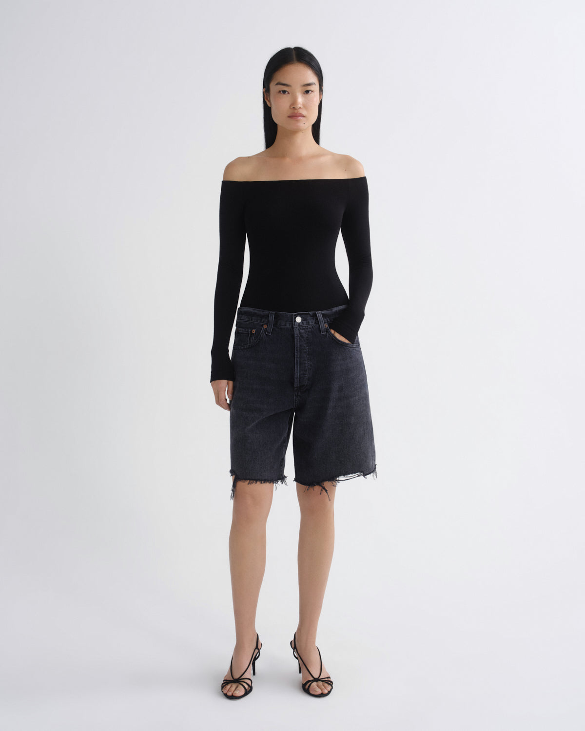A model stands against a light gray studio background wearing a black off-the-shoulder long-sleeve top, dark gray denim shorts with a raw hem, and black strappy high-heeled sandals.