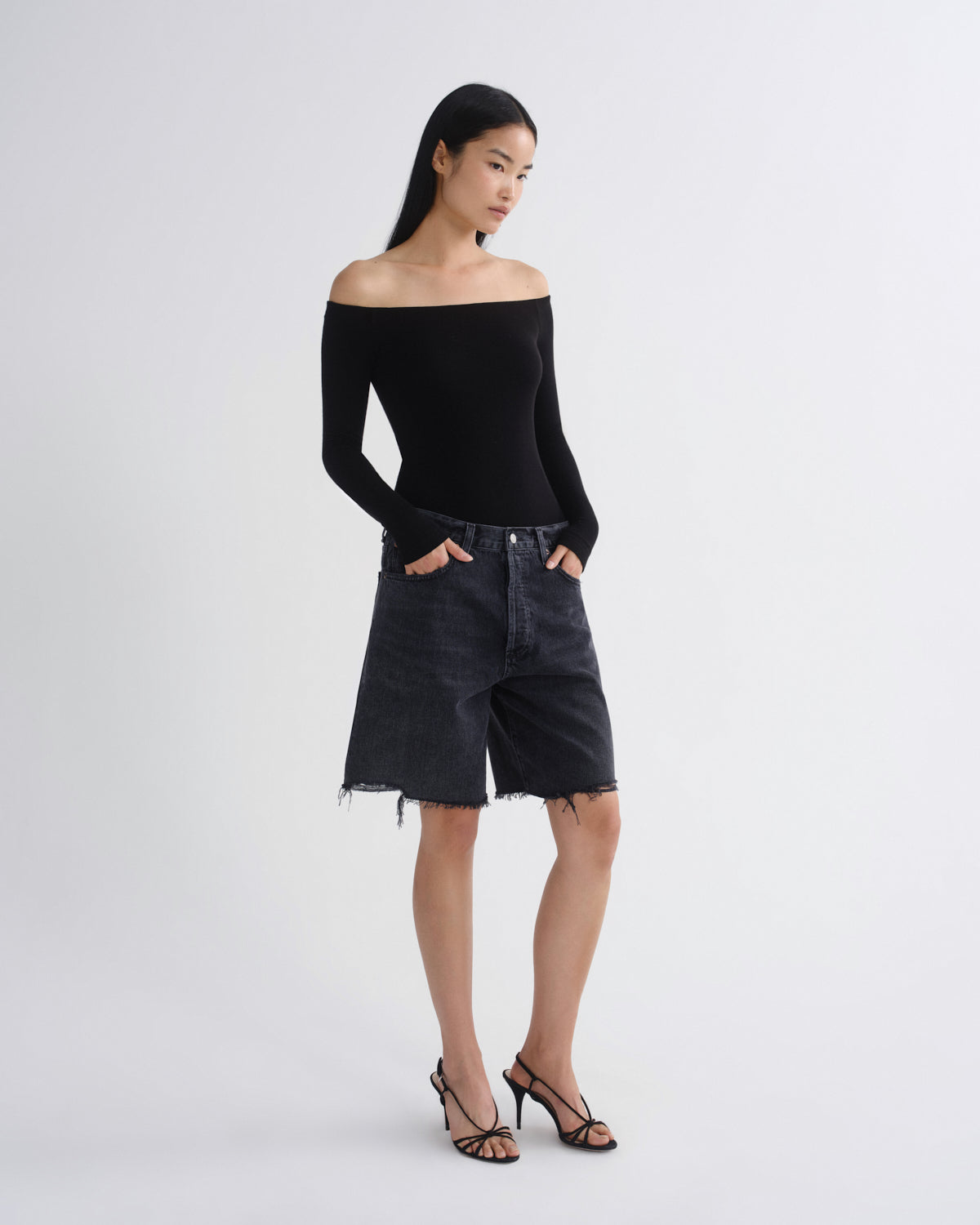 A model stands against a light gray studio background wearing a black off-the-shoulder long-sleeve top, dark washed denim shorts with raw hems, and black strappy high-heeled sandals.