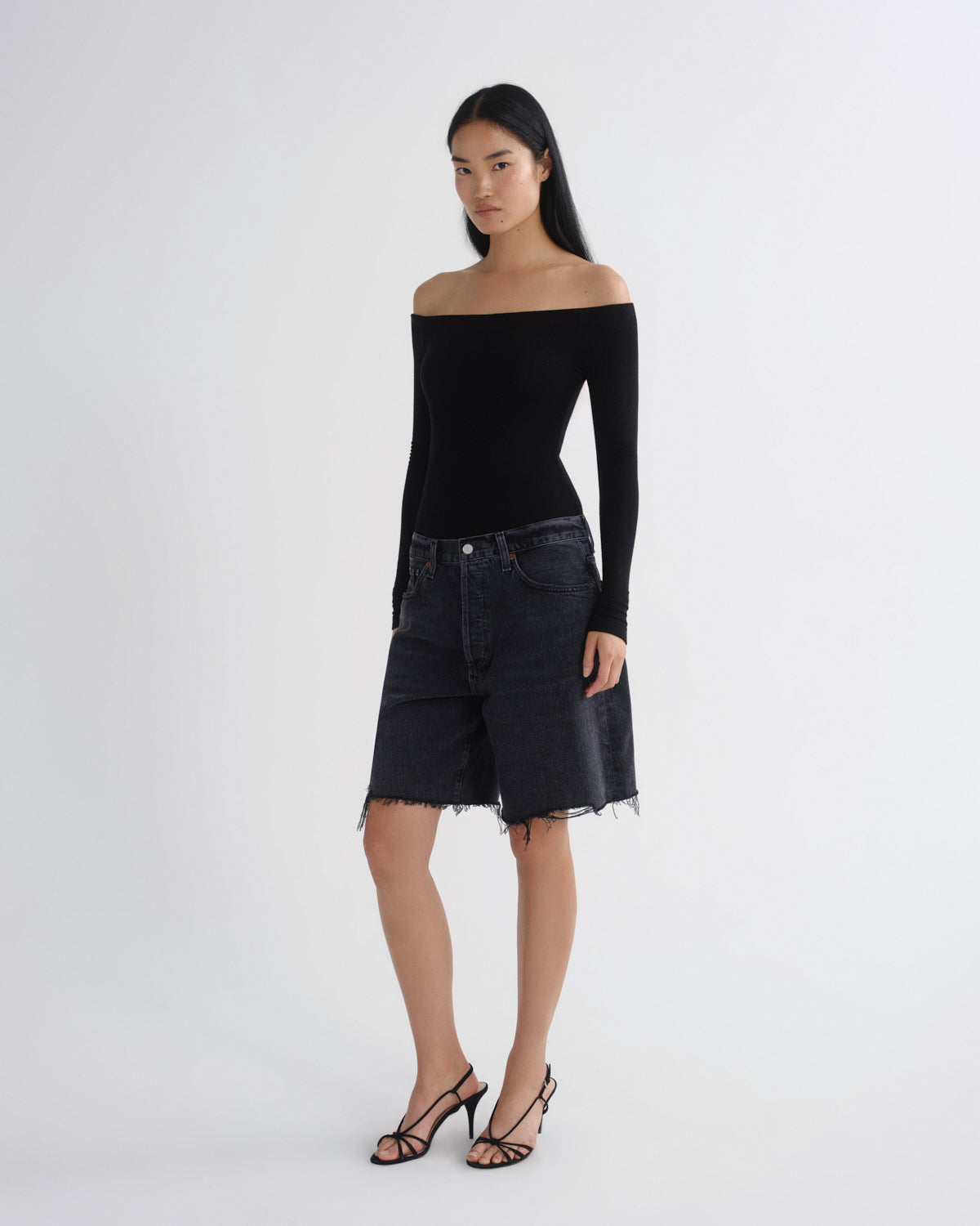 A model stands against a light gray studio background wearing a fitted black off-the-shoulder long-sleeve top, dark washed denim shorts with a raw hem, and black strappy high-heeled sandals.