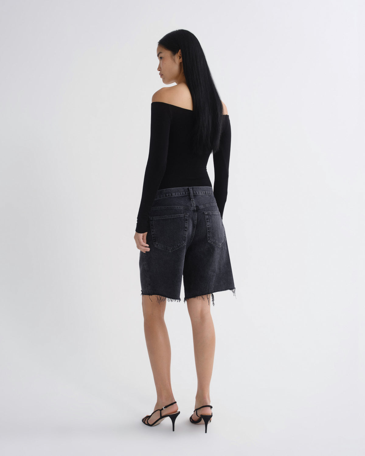 A woman is shown from behind wearing a black off-the-shoulder long-sleeve top, black denim shorts with a raw hem, and black strappy high-heeled sandals against a plain white studio background.