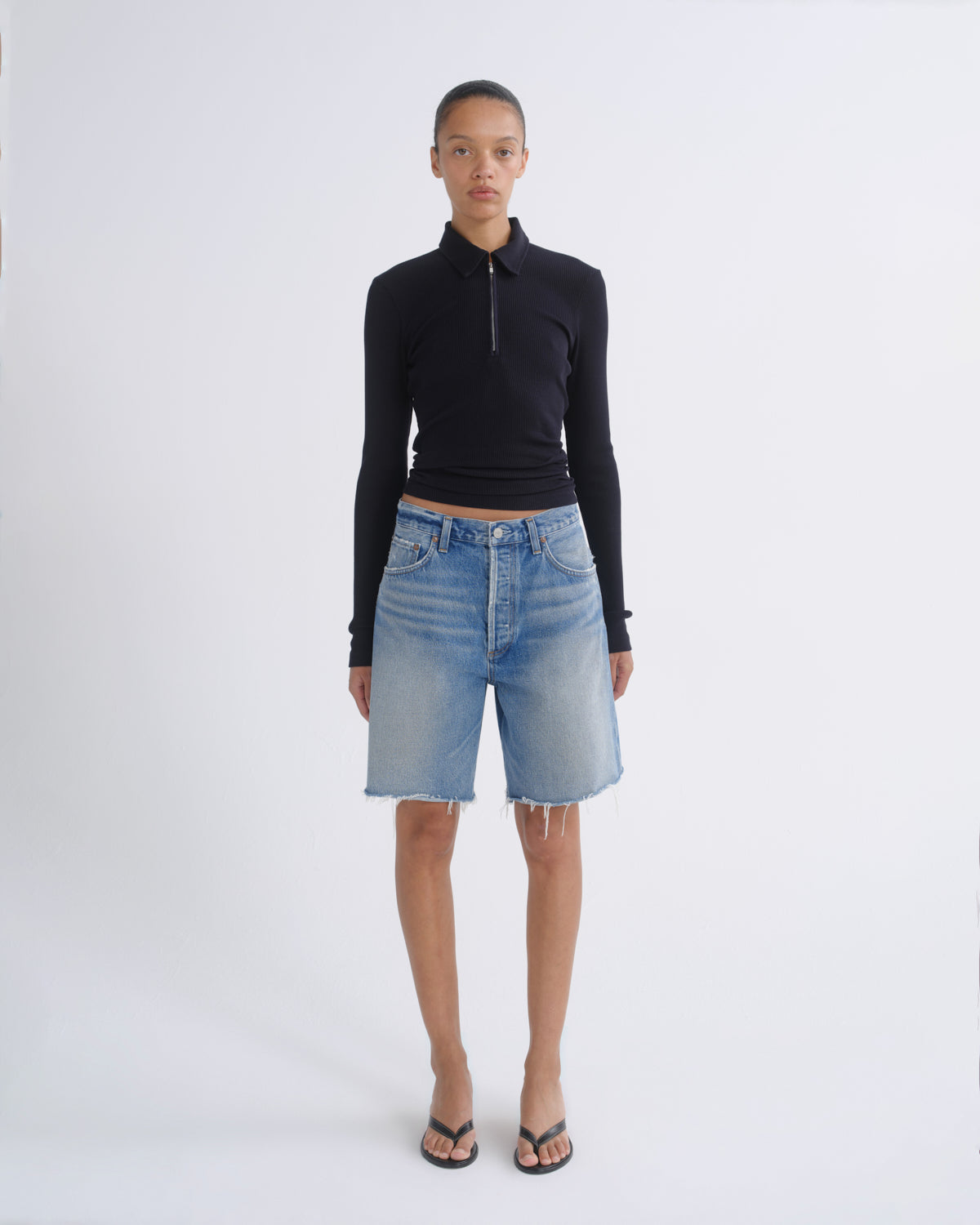 A model stands against a light gray studio background wearing a black long-sleeve half-zip polo top, light-wash blue denim shorts, and black flip-flop sandals.