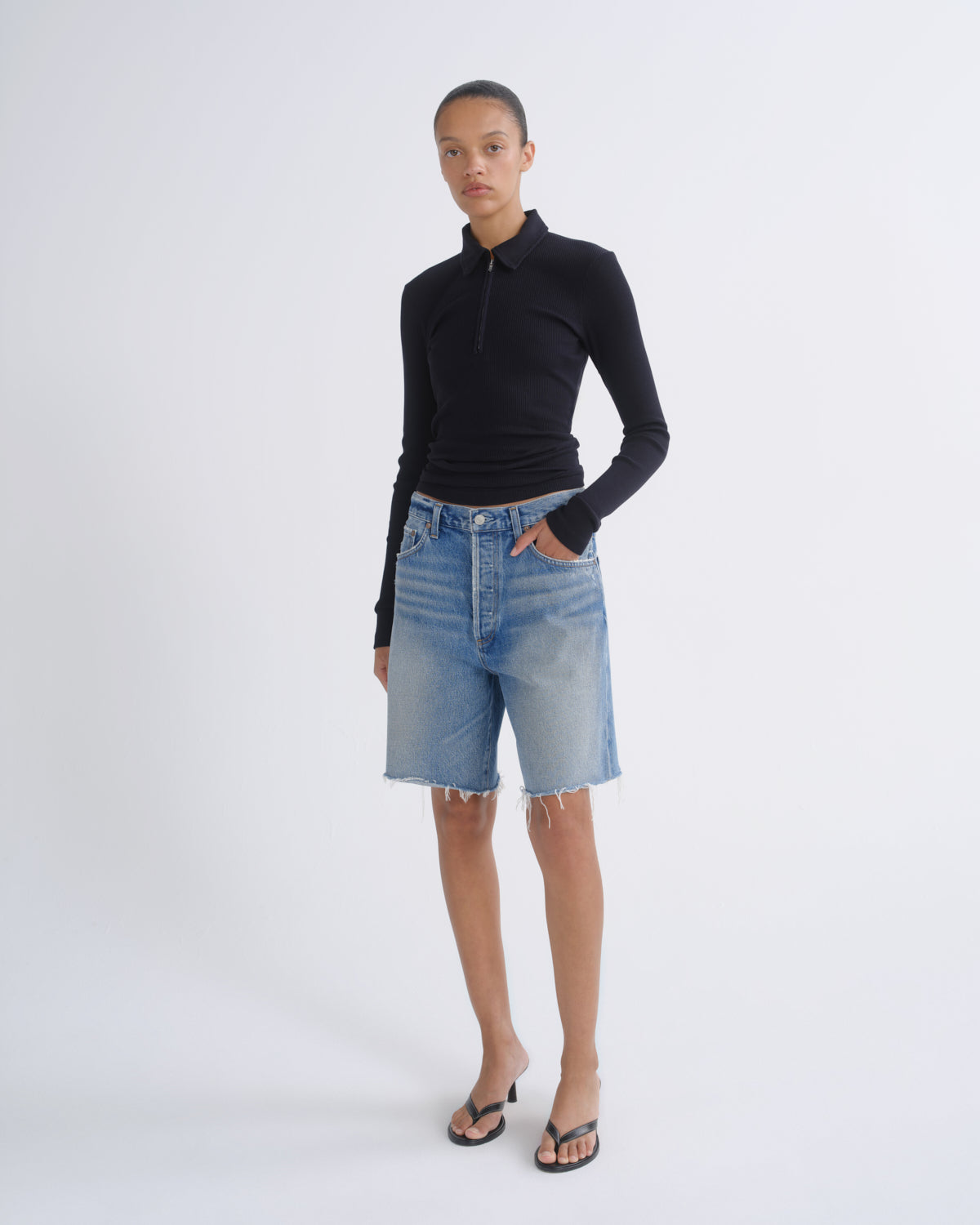 A model stands against a light gray studio background wearing a fitted black long-sleeve polo shirt, light-wash blue denim shorts with raw hems, and black thong sandals.