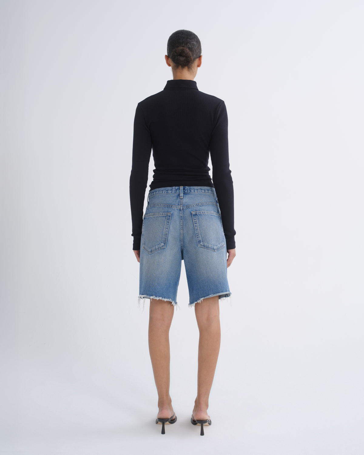A model shown from the back wearing a black long-sleeve mock neck top, light blue denim shorts, and black high-heeled sandals, set against a plain light gray studio background.