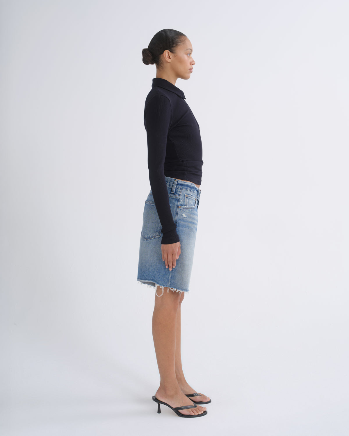 A woman stands in profile wearing a fitted black long-sleeve top, a light blue denim mini skirt, and black heeled sandals against a plain light gray studio background.