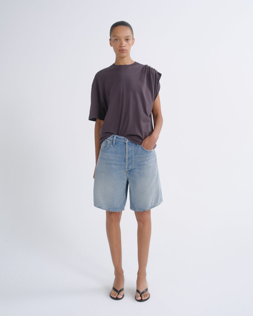 A model stands against a light gray studio background wearing a dark plum-purple asymmetrical short-sleeve t-shirt, light-wash blue denim shorts, and black flip-flop sandals.