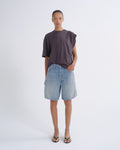 A model stands against a light gray studio background wearing a dark plum-purple asymmetrical short-sleeve t-shirt, light-wash blue denim shorts, and black flip-flop sandals.