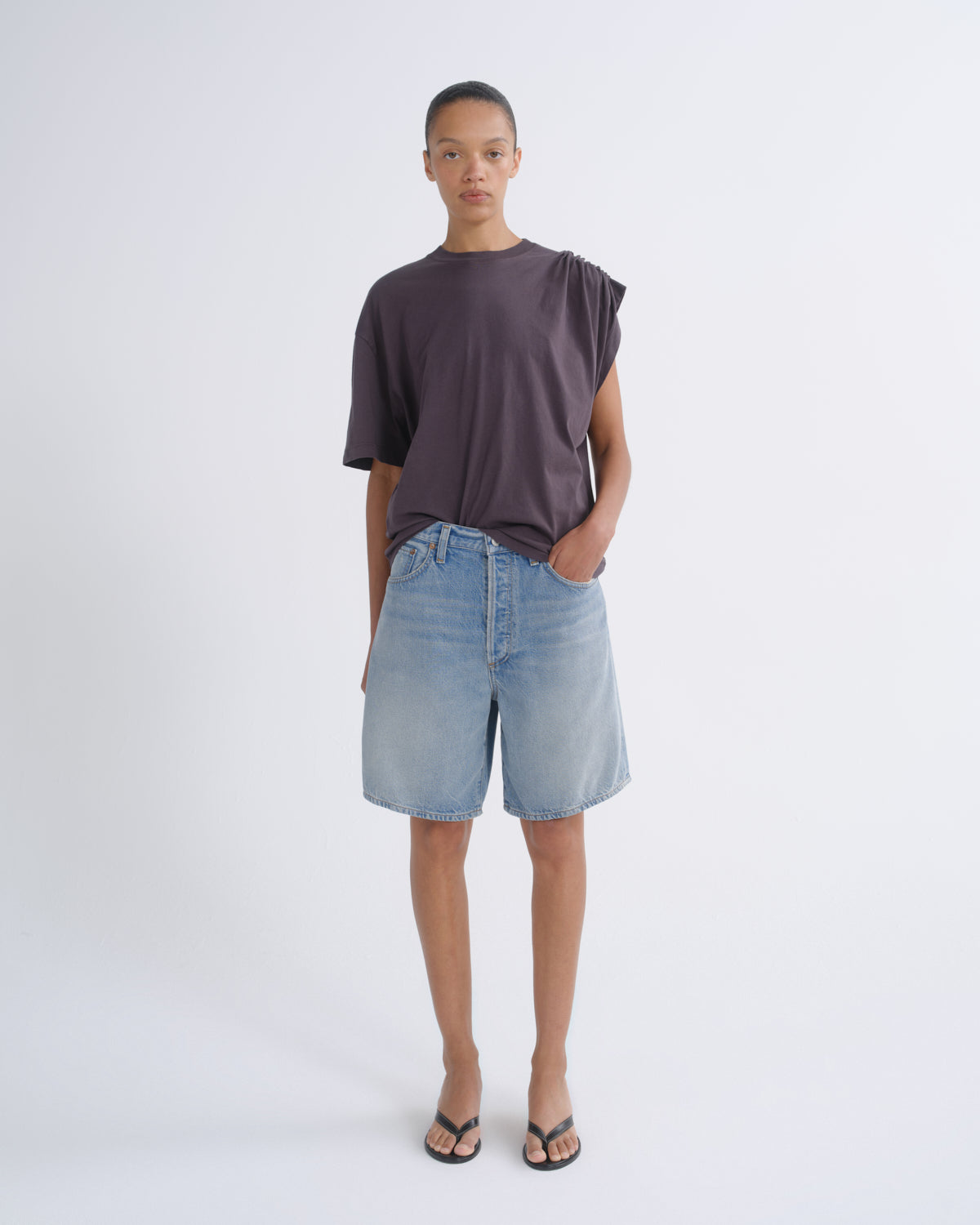 A model stands against a light gray studio background wearing a dark plum-purple asymmetrical short-sleeve t-shirt, light-wash blue denim shorts, and black flip-flop sandals.