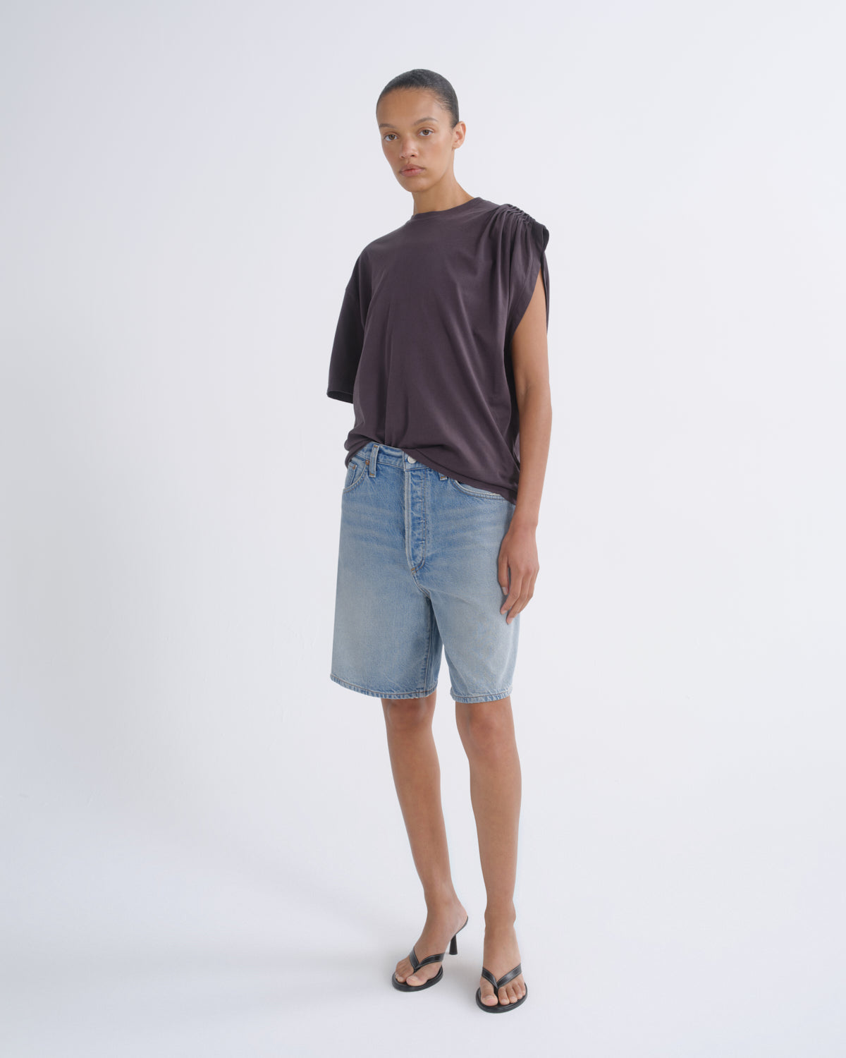 A model stands against a light gray studio background wearing a dark plum-purple draped short-sleeve top, light-wash denim shorts, and black heeled sandals.