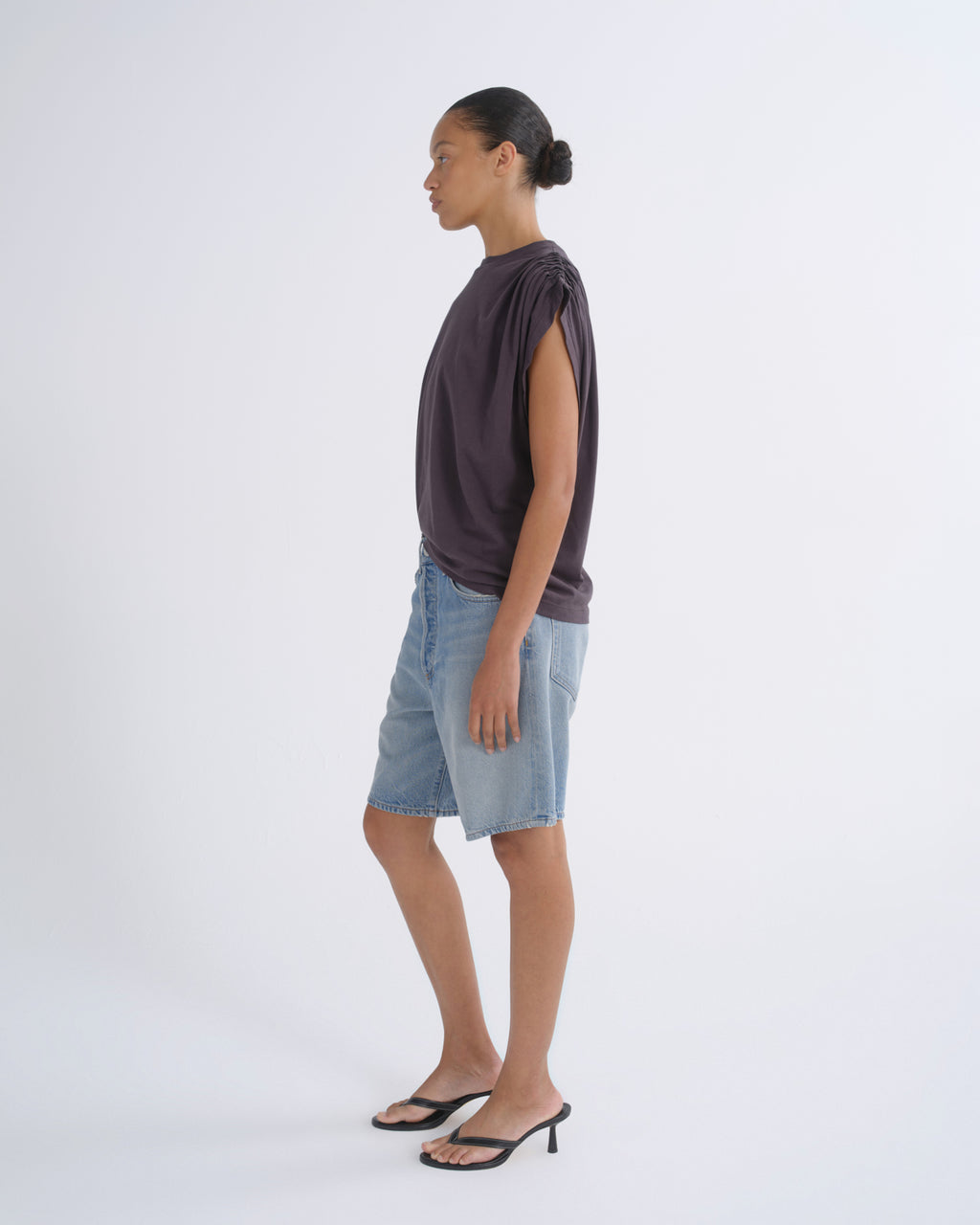 A model shown in profile wearing a dark plum-purple sleeveless top, light-wash denim shorts, and black heeled sandals against a clean white studio background.