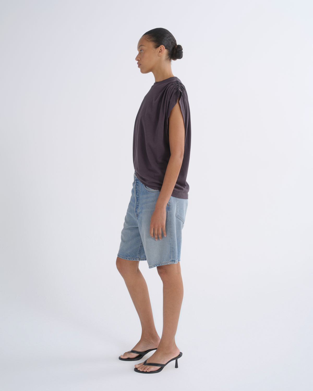 A model shown in profile wearing a dark plum-purple sleeveless top, light-wash denim shorts, and black heeled sandals against a clean white studio background.