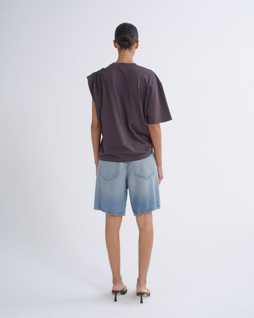 A model viewed from behind wearing a dark plum-purple oversized short-sleeve t-shirt and light blue denim shorts, paired with black high-heeled sandals, against a plain light gray studio background.