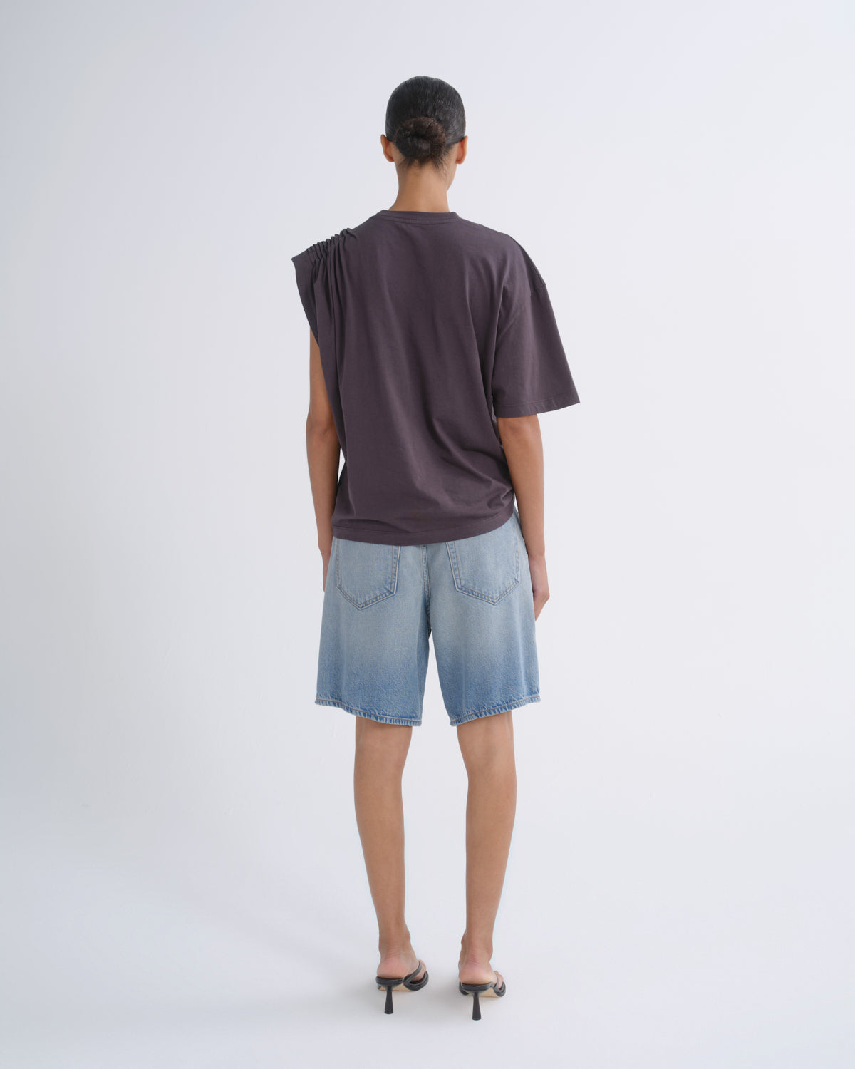 A model viewed from behind wearing a dark plum-purple oversized short-sleeve t-shirt and light blue denim shorts, paired with black high-heeled sandals, against a plain light gray studio background.