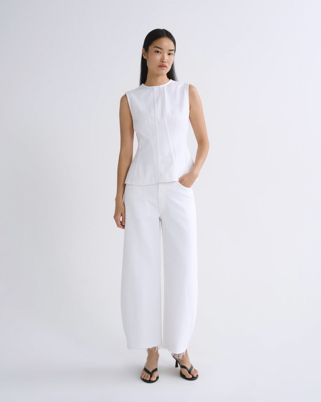 A model stands against a plain light gray studio background wearing a white sleeveless top and matching white wide-leg pants, paired with black heeled sandals.
