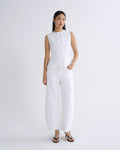 A model stands against a plain light gray studio background wearing a white sleeveless top and matching white wide-leg pants, paired with black heeled sandals.