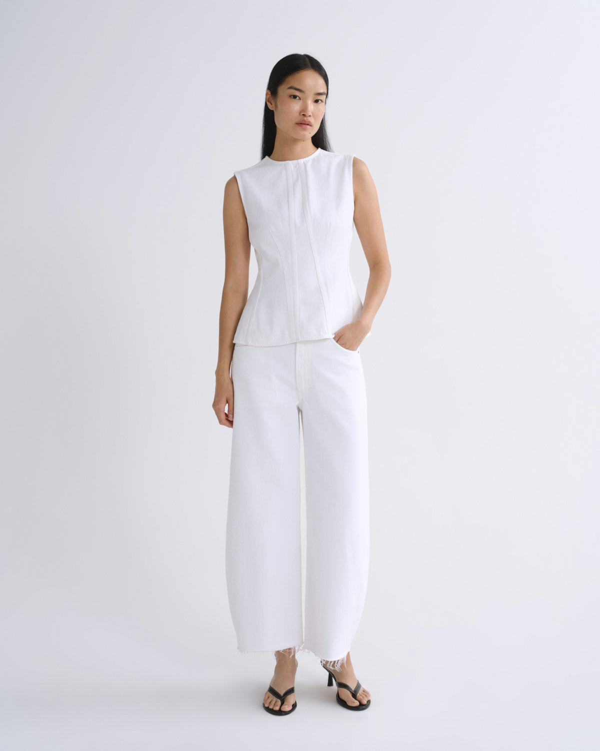A model stands against a plain light gray studio background wearing a white sleeveless top and matching white wide-leg pants, paired with black heeled sandals.
