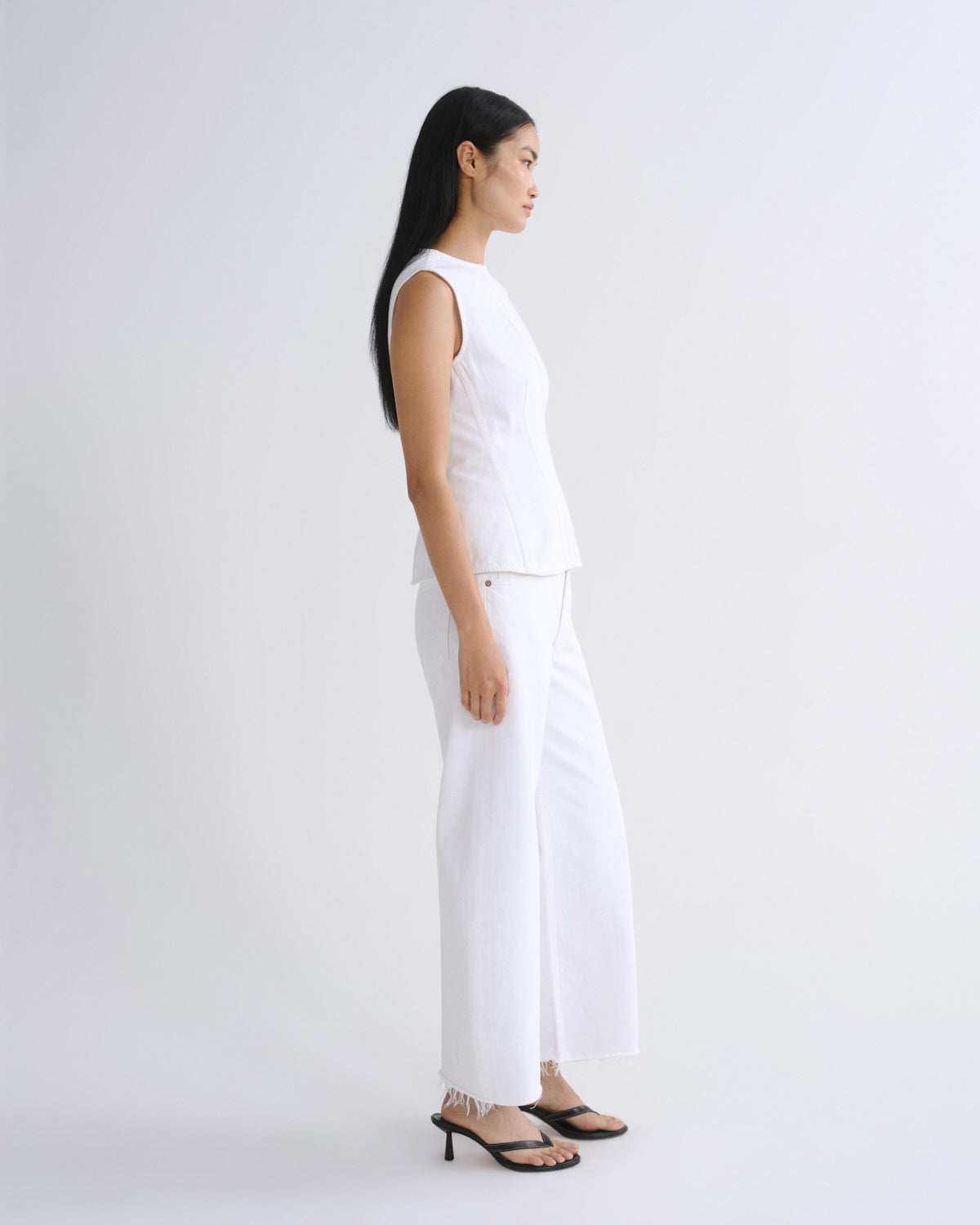 A woman stands in profile wearing a white sleeveless top and matching white wide-leg pants, paired with black heeled sandals, against a plain light gray studio background.