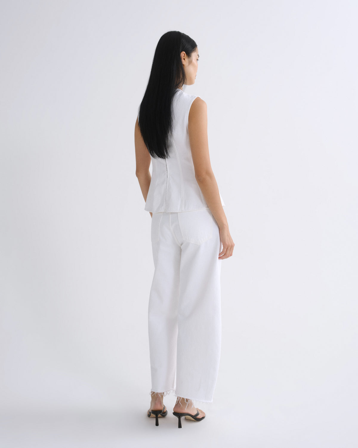 A woman with long black hair is shown from the back wearing a white sleeveless top and matching white wide-leg trousers, paired with black high-heeled sandals, against a plain white studio background.