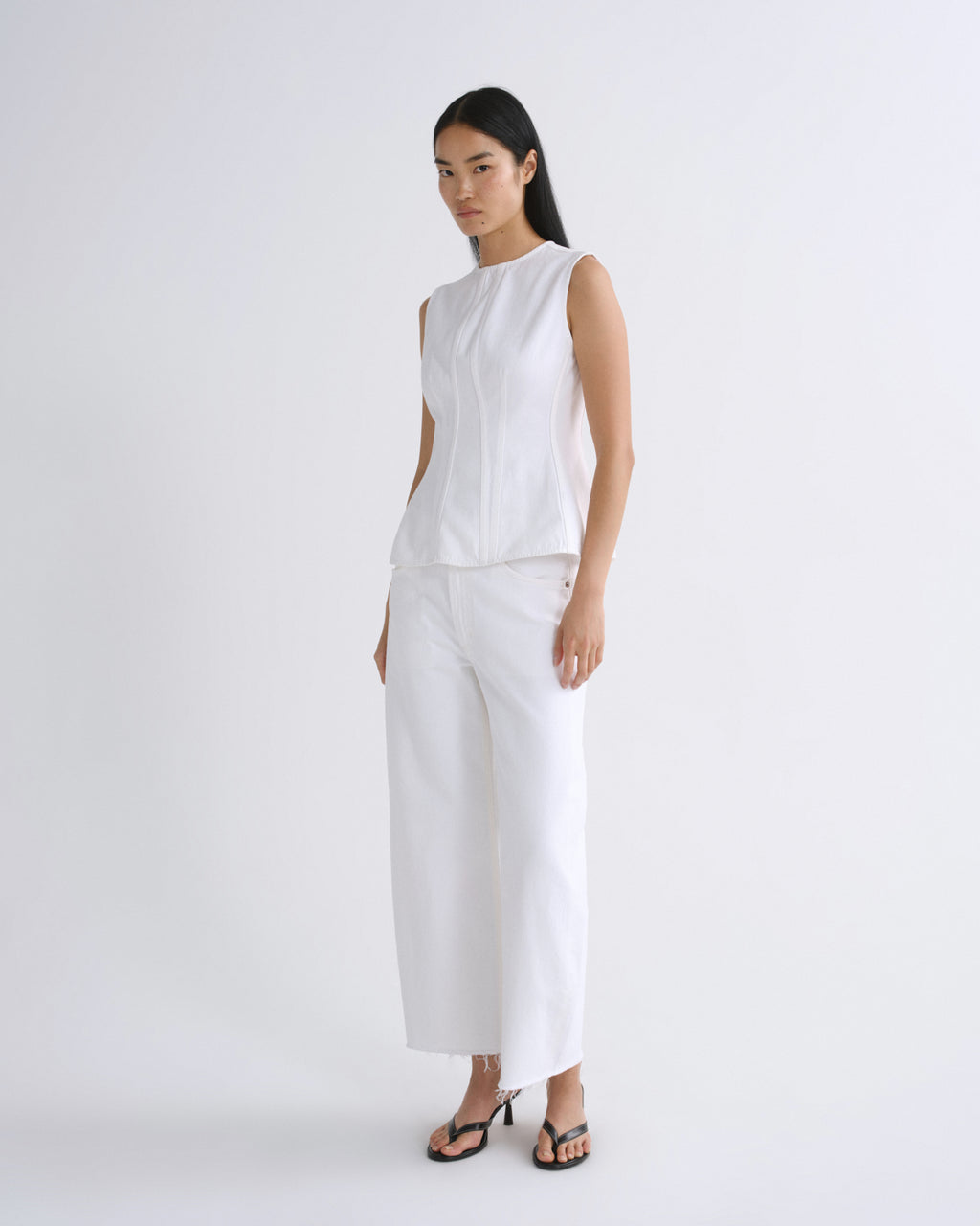 A model wearing a white sleeveless top and matching white wide-leg cropped pants, paired with black strappy heels, standing against a plain white studio background.