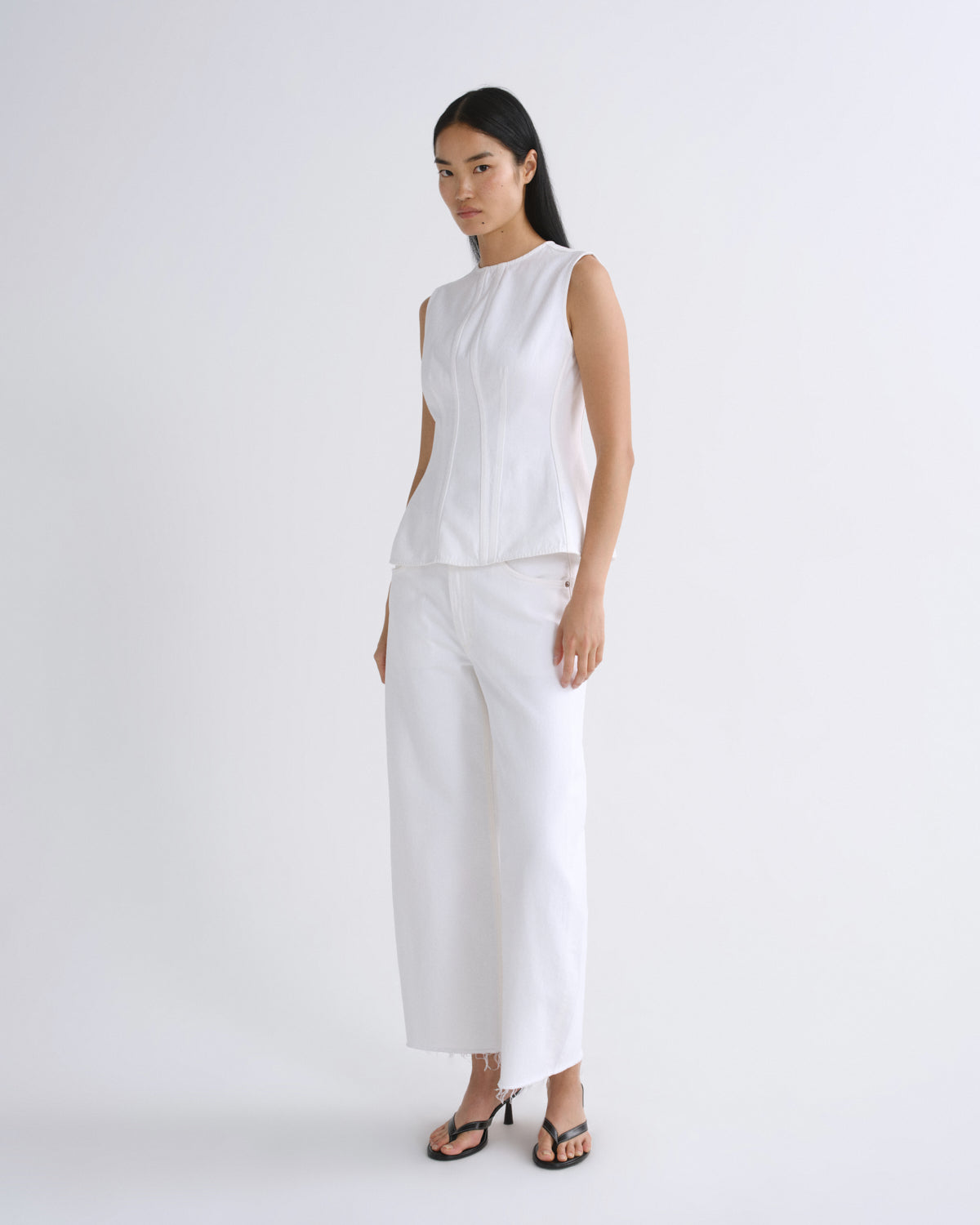 A model wearing a white sleeveless top and matching white wide-leg cropped pants, paired with black strappy heels, standing against a plain white studio background.