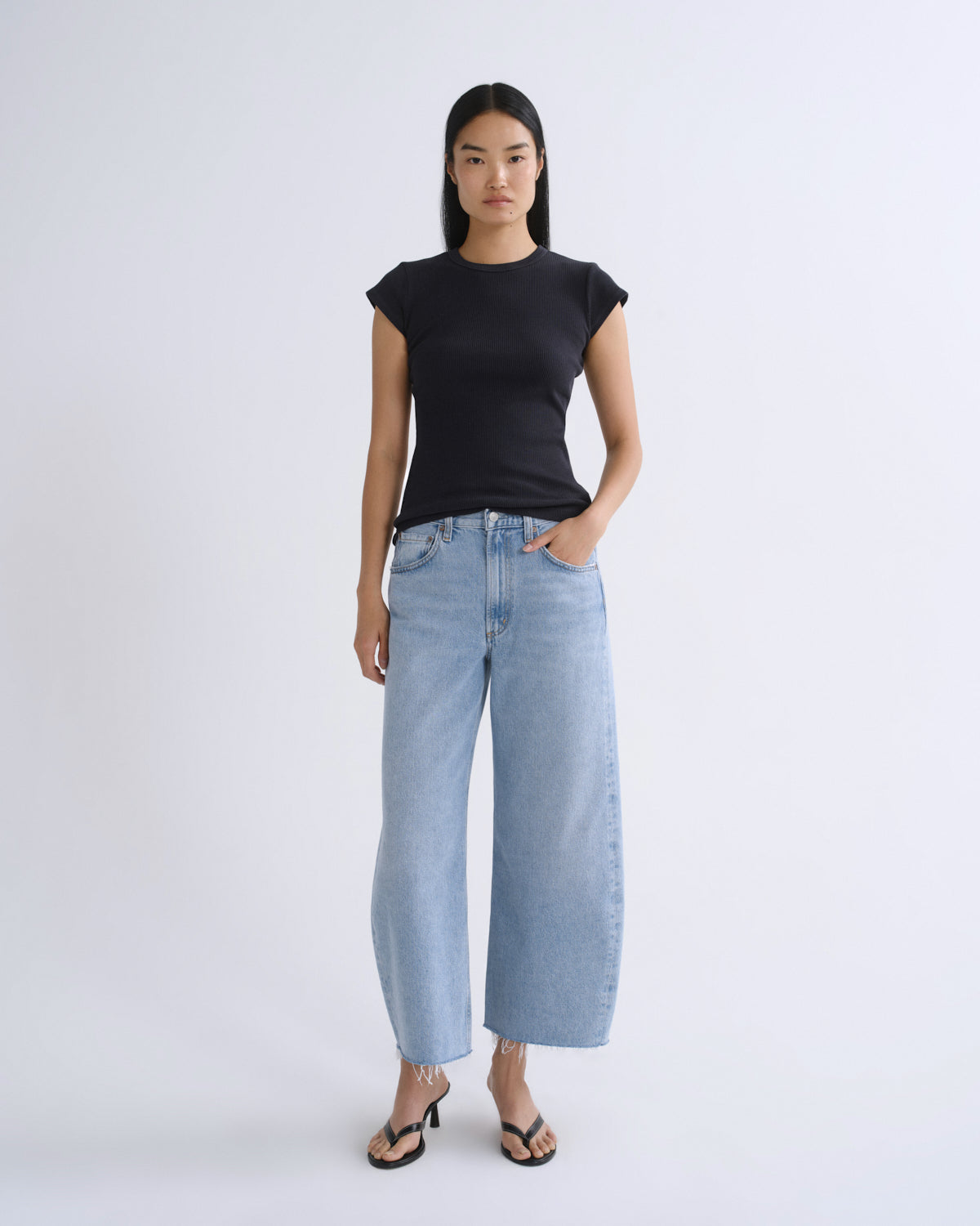 A model stands against a light gray studio background wearing a black short-sleeve crewneck t-shirt, light-wash wide-leg cropped jeans, and black heeled sandals.