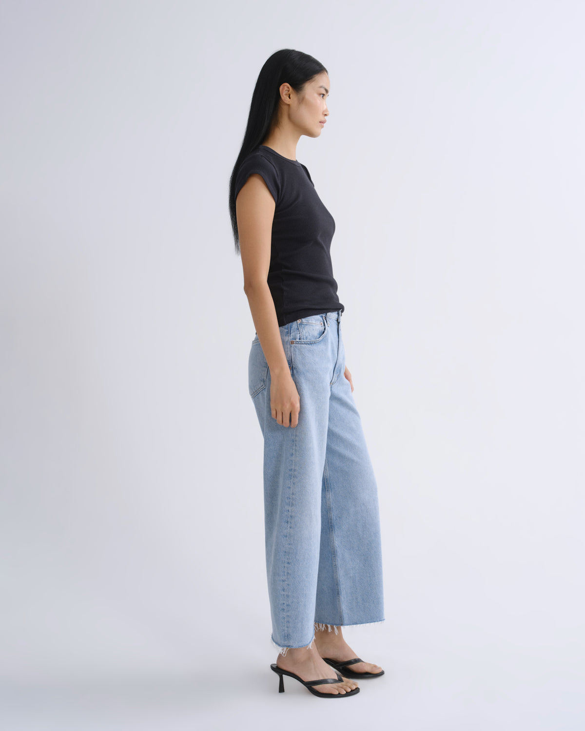 A woman in a black sleeveless top and light blue wide-leg jeans stands in profile against a plain light gray studio background.